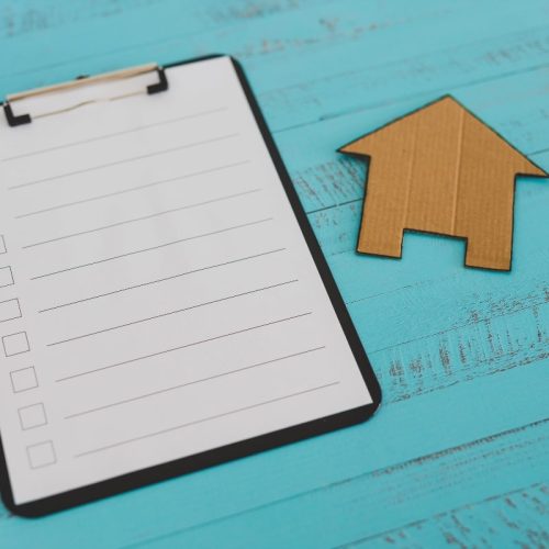 A blank checklist with checkboxes, a pencil, and a cardboard house placed on a turquoise wooden background, symbolising a list of things to do when moving house with a focus on home security.